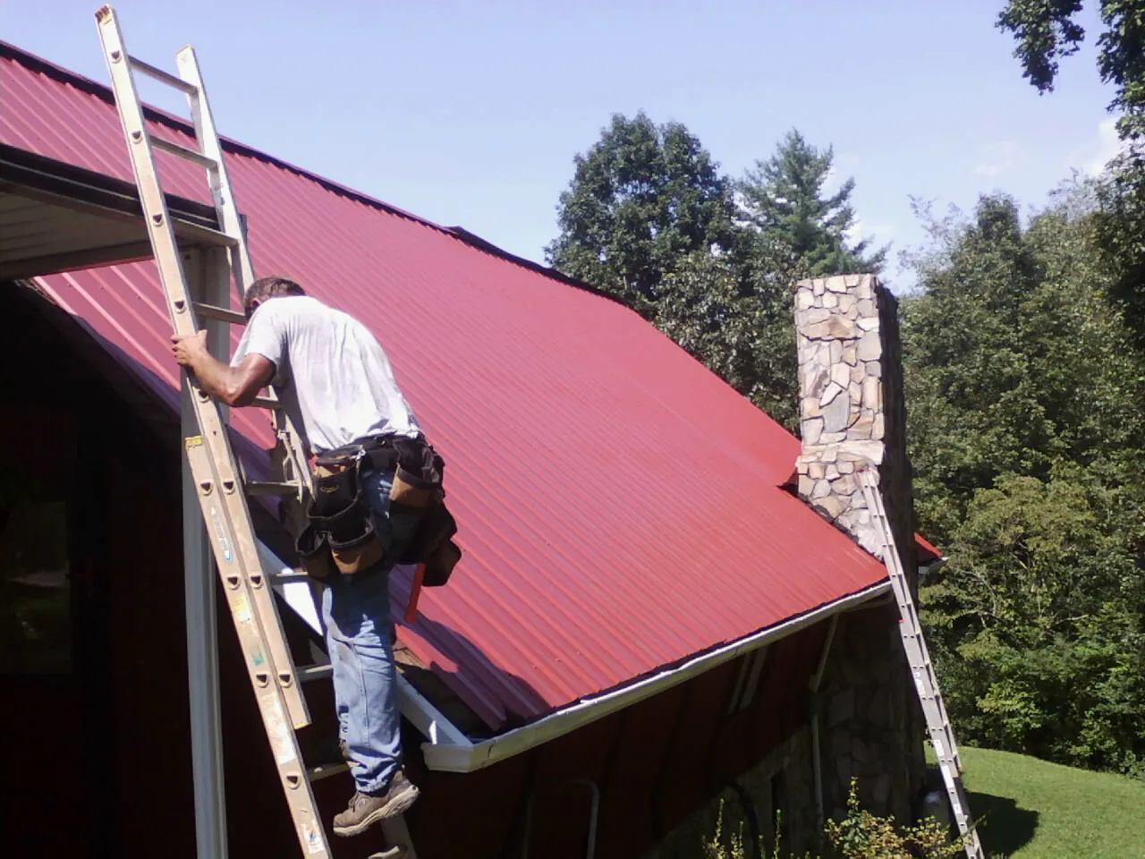 Roofer on a ladder beside a red metal roof and stone chimney — Soffit & Fascia Repair by Roofing Brothers in Tiro